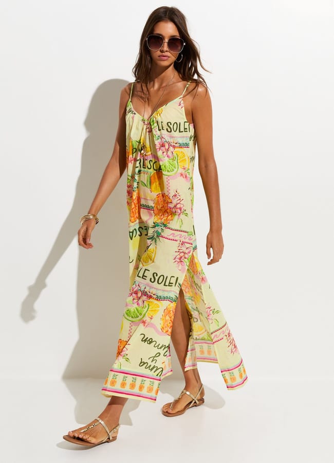 A woman wearing sunglasses and a colourful, sleeveless maxi dress with vibrant fruit prints and French text stands against a white wall. She is wearing sandals and jewellery, and her hair is loose.