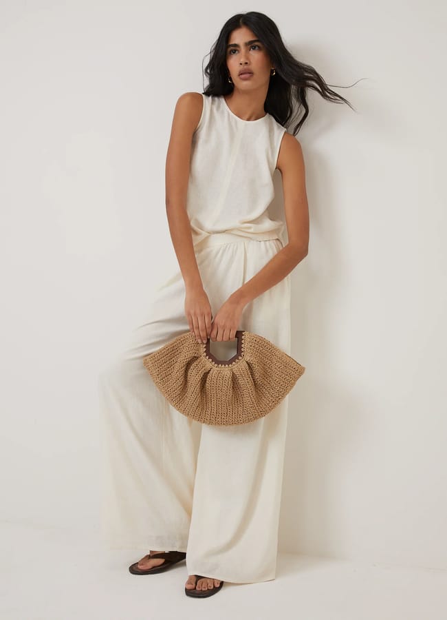 Woman wearing a high street linen off-white sleeveless top and wide-leg trousers stands against a white wall, holding a woven tan handbag and wearing dark flat sandals. Her long dark hair is loose, and she has a relaxed pose.