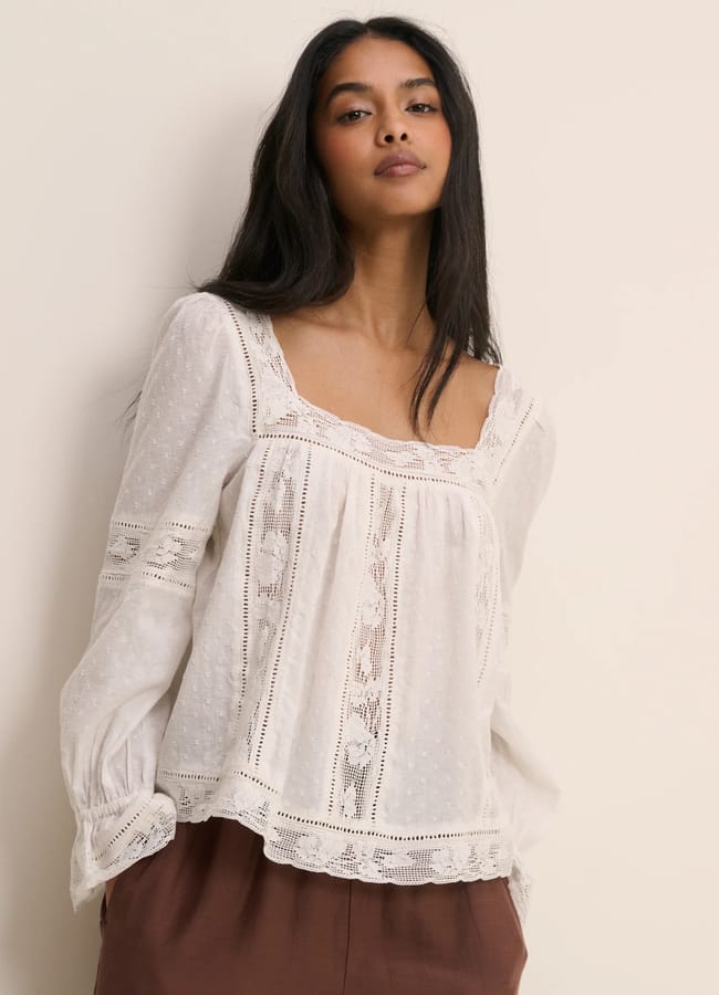 A woman with long dark hair wears a loose, white, long-sleeved blouse with lace details and brown trousers, standing against a plain light-coloured background—an effortlessly chic look perfect for browsing Father’s Day gifts 2026.
