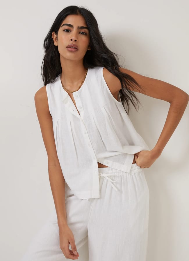 A woman with long dark hair wears a sleeveless high street linen button-up top and matching white trousers, standing against a plain light background with a relaxed pose and neutral expression.