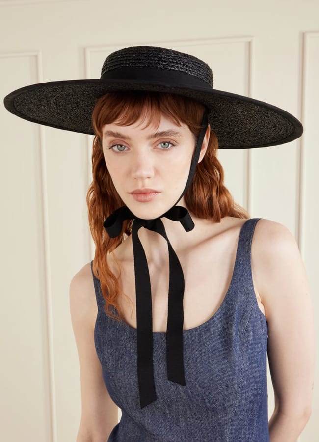 A woman with light skin and long red hair wears a wide-brimmed black hat inspired by modern fascinator trends, tied with a ribbon under her chin, and a sleeveless blue denim dress, standing before a cream-coloured panelled wall.