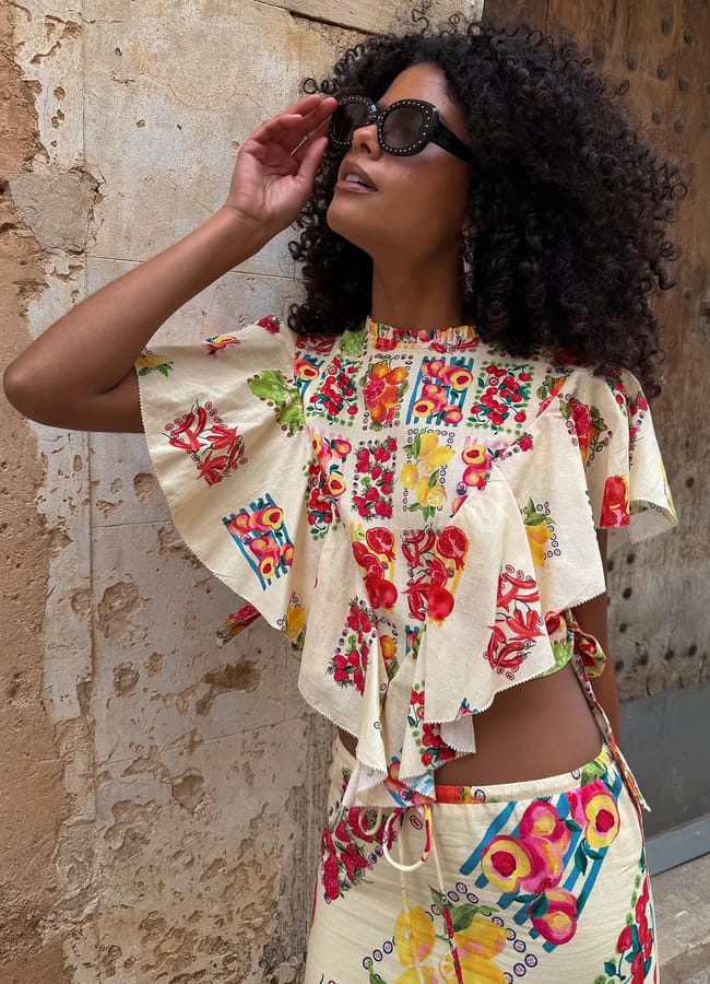 A woman with curly hair and sunglasses poses against a textured wall, wearing a colourful, fruit-print top with ruffled sleeves and a matching skirt.