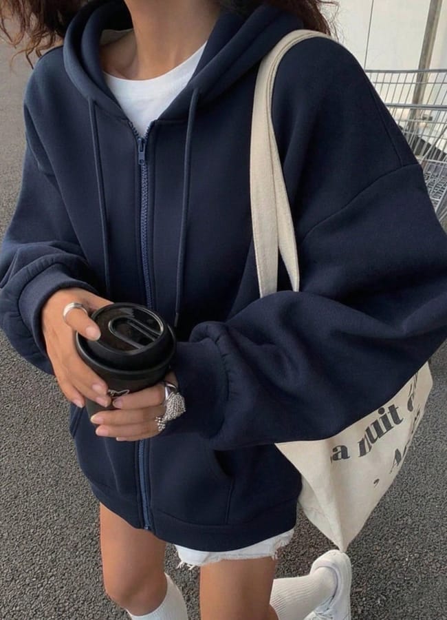 A person wearing an oversized navy blue zip-up hoodie, white shorts, and knee-high socks gives off serious cute padel outfits vibes. They hold a black travel mug and a cream-coloured tote bag, with a shopping trolley in the background.