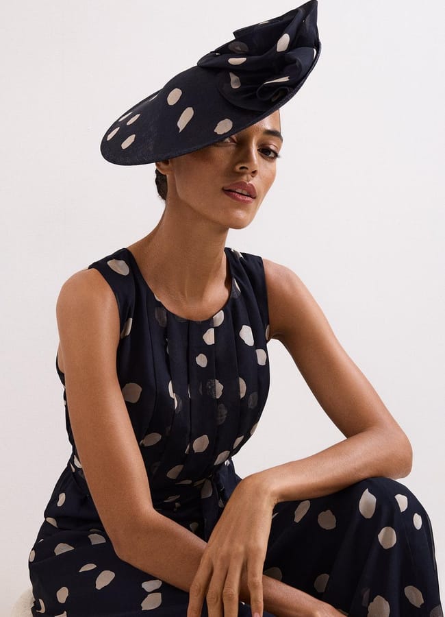 A woman wearing a sleeveless navy dress with large beige polka dots and a matching wide-brimmed hat—giving her own hot take on fashion accessories—poses confidently against a plain, light background.