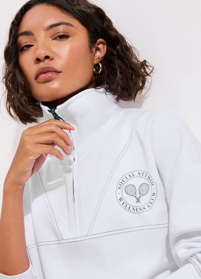 A woman with wavy brown hair wears a white zip-up sweatshirt featuring a round emblem with two tennis racquets and the text “Social Studio Wellness Club.” She gazes confidently at the camera, hand near her chin—serving inspiration for cute padel outfits.