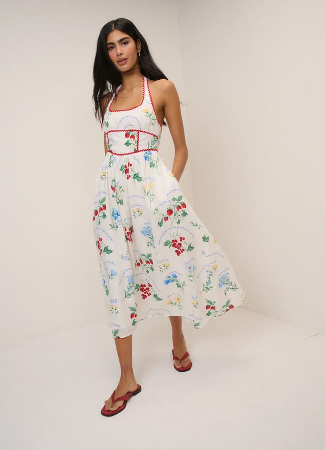 A woman stands in a simple room, wearing a sleeveless white midi dress adorned with colourful fruit prints and red trim, paired with red sandals. She has long dark hair and a relaxed pose.