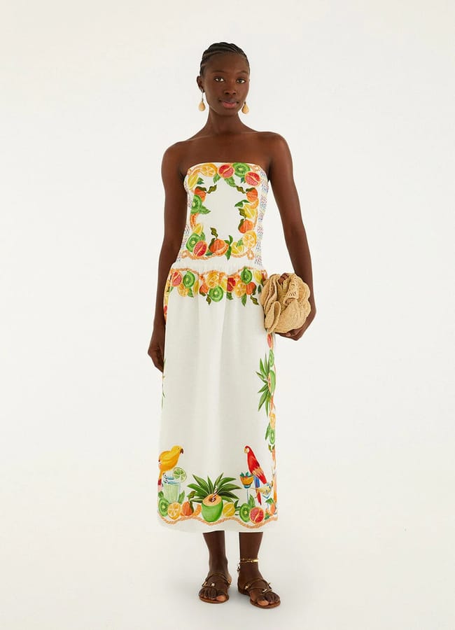 A woman wearing a strapless white dress with vibrant fruit prints and bird patterns, holding a tan woven clutch, and accessorised with sandals and gold earrings, stands against a plain light background.