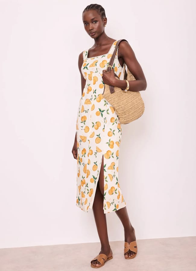 A woman wearing a sleeveless, white midi dress with playful fruit prints and a side slit stands against a plain background. She carries a woven straw bag and wears brown sandals and gold jewellery.