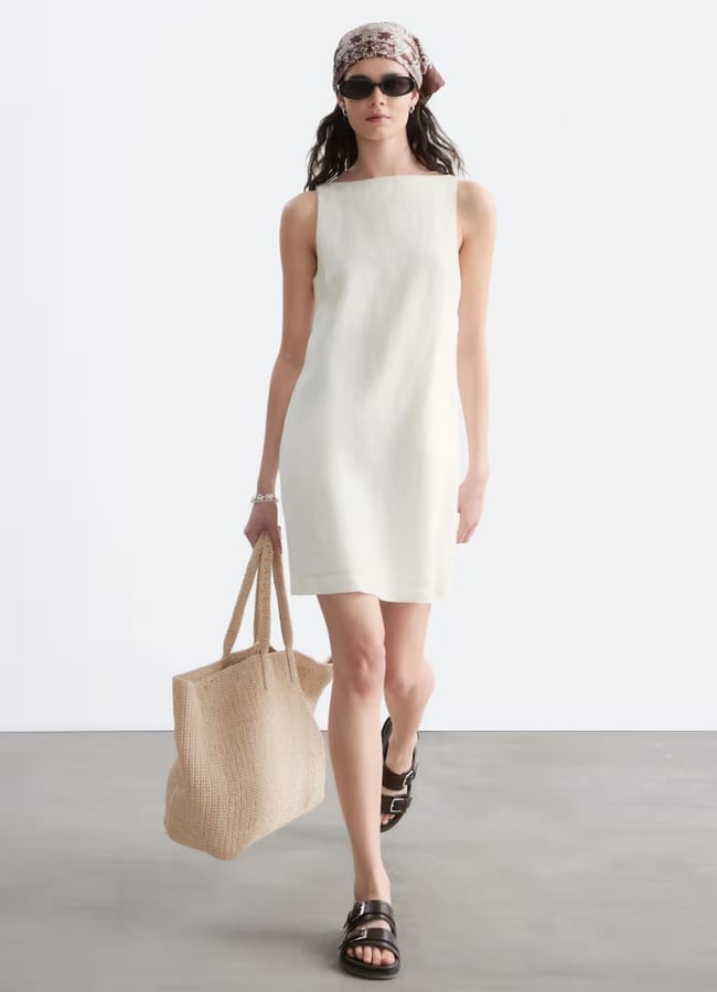 A woman wearing a high street linen sleeveless white dress, sandals, a patterned headscarf, and sunglasses carries a large woven tote bag whilst walking indoors.