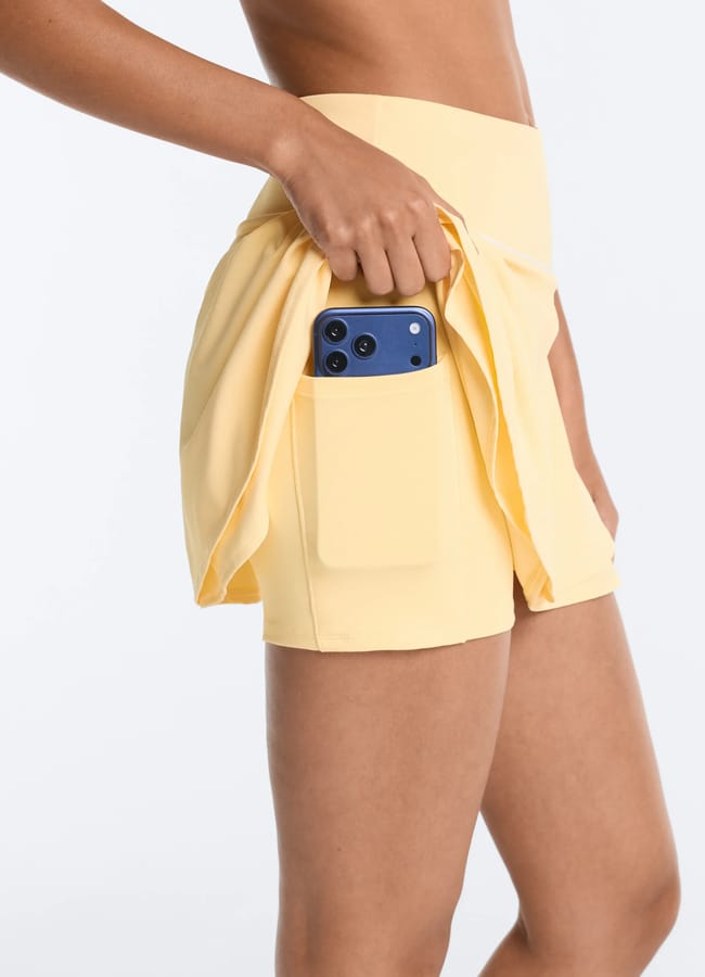 A person wearing yellow athletic shorts, ideal for cute padel outfits, places a blue mobile phone into a side pocket on the shorts. The background is plain white.