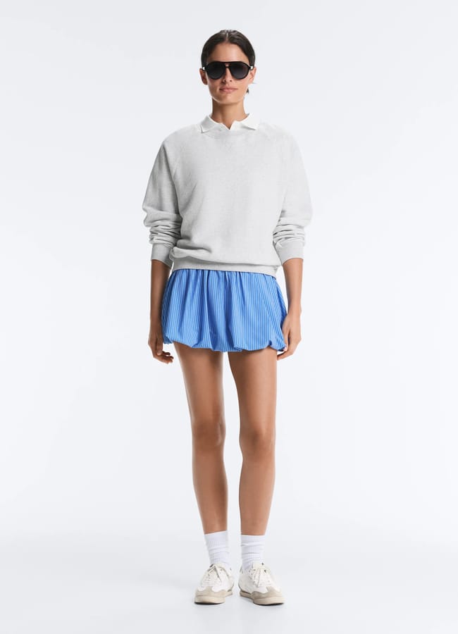 A person wearing sunglasses, a light grey jumper, a collared shirt, a blue pleated skirt, white socks, and beige trainers stands against a plain white background—showcasing one of the cute padel outfits perfect for on or off the court.