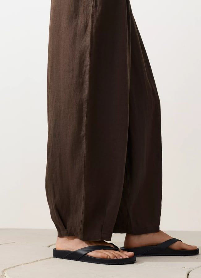 A person wearing loose, brown, high street linen wide-leg trousers and black sandals stands on a light-coloured surface against a plain background. Only the lower legs and feet are visible.