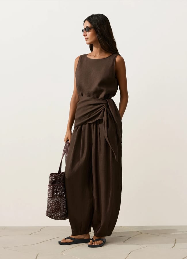 Woman wearing a sleeveless brown high street linen top with a side tie, matching wide-leg trousers, black sandals, sunglasses, and carrying a patterned tote bag, standing against a plain light background.