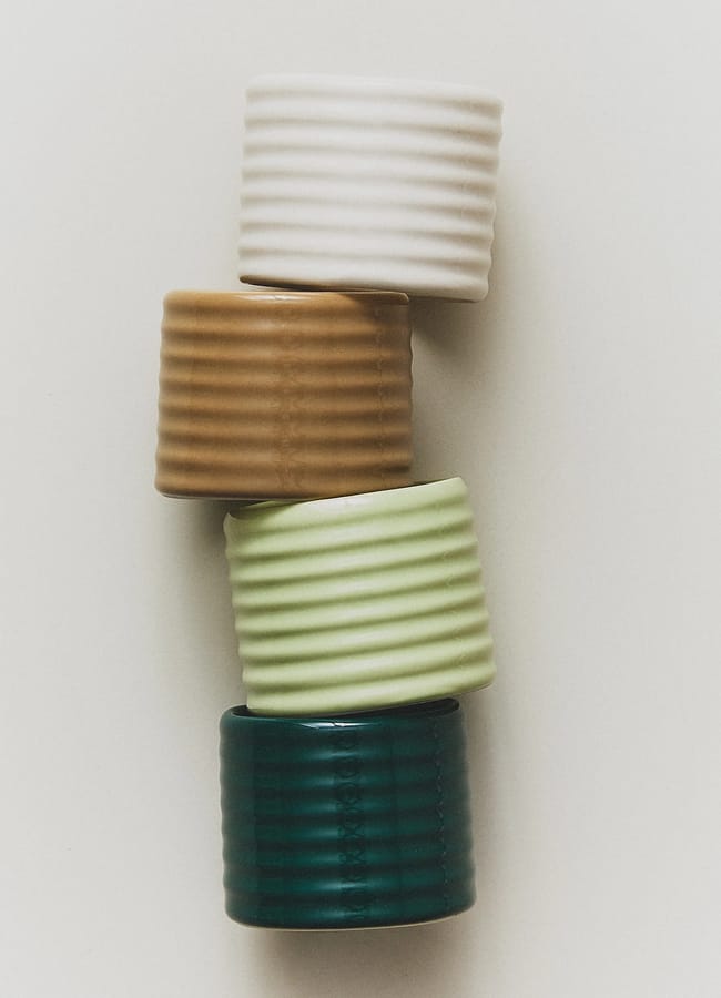 Four ribbed ceramic cups in white, brown, light green, and dark green are stacked vertically against a plain white background—perfect for those seeking affordable interiors with a stylish touch.