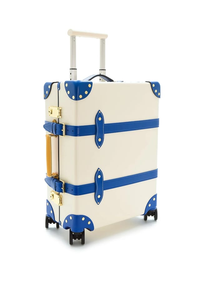 A cream-coloured hard-shell suitcase from one of the best suitcase brands, featuring blue leather straps and accents, gold fittings, a tan side handle, telescopic handle, and four black wheels, standing upright on a white background.