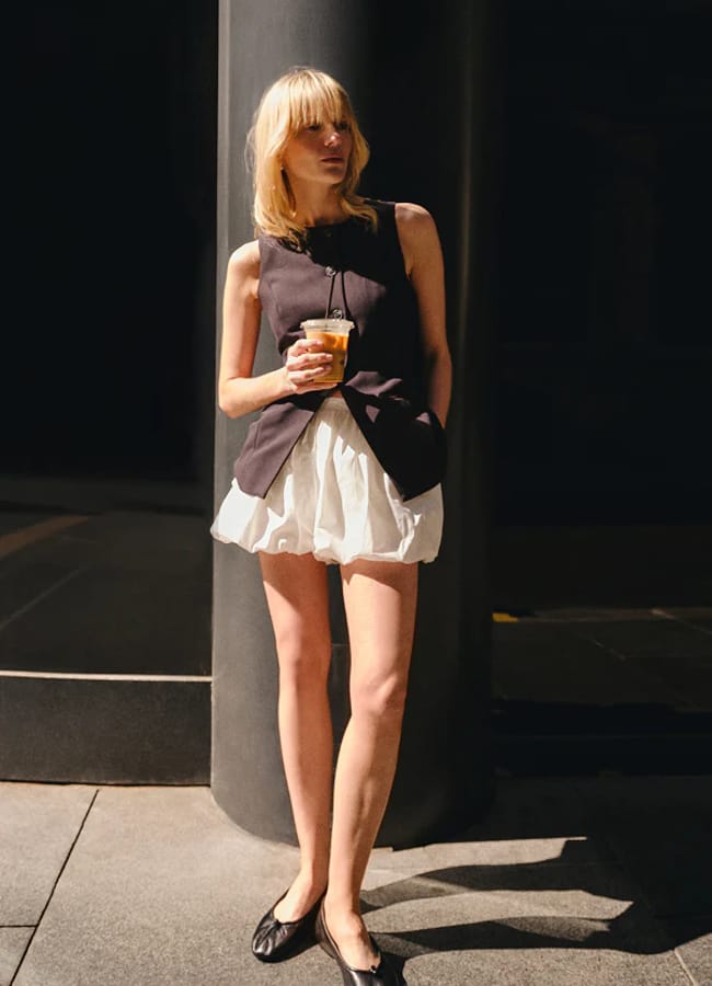 A person with blonde hair stands on a city pavement at night, holding a drink. They wear a black sleeveless top, white bubble shorts, and black ballet pumps, with one hand in their pocket.
