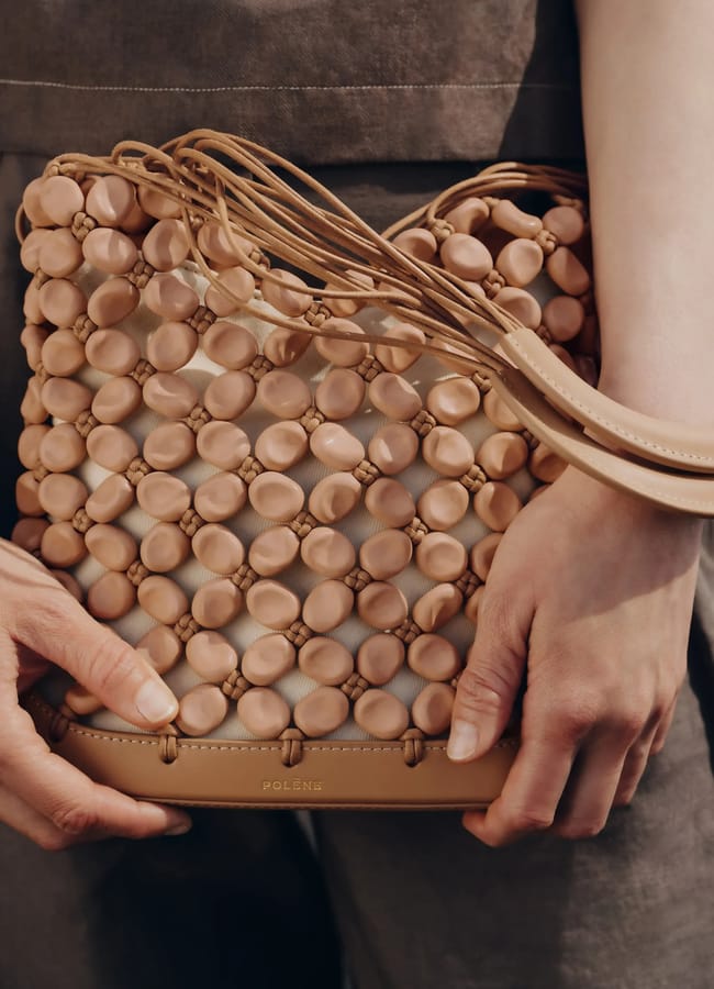 A person holds a stylish tan beaded bag made of interconnected round beads and leather, with a plaited strap. The brand name Polène is visible on the bottom edge of the bag.