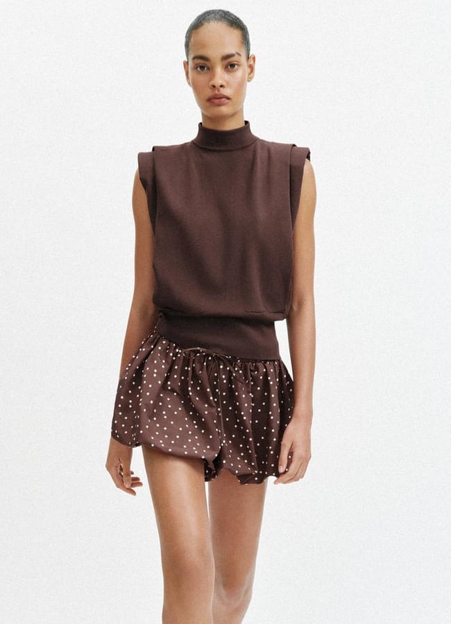A woman wearing a sleeveless brown polo-neck top and brown sheer polka-dot bubble shorts walks forward against a plain white background.