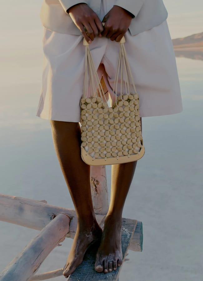 A person with dark skin stands barefoot on a wooden log, wearing a light-coloured dress and holding a beaded bag crafted from round beads. The scene is serene, set against a calm, water-like background.