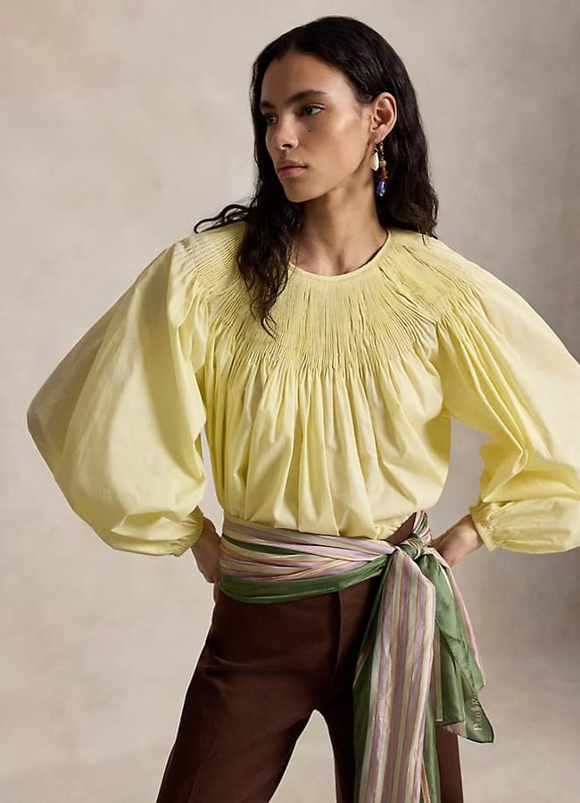 A woman stands with hands on hips, wearing a yellow puff-sleeve blouse with gathered detailing, brown trousers, and a pastel striped sash tied at the waist—an effortlessly stylish look perfect for those seeking unique Fathers’ Day gifts 2026.