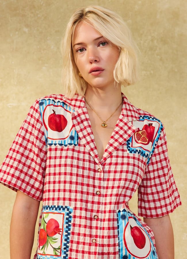A woman with blonde hair wearing a red and white chequered shirt adorned with fruit prints, including pomegranate illustrations, stands against a beige background.