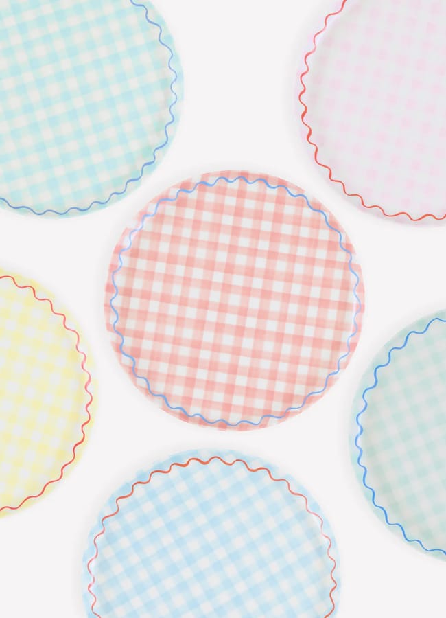 Six round plates with pastel gingham patterns in blue, pink, and yellow, each with a wavy coloured edge, are arranged in a scattered pattern on a white background—perfect picnic essentials for any outdoor gathering.