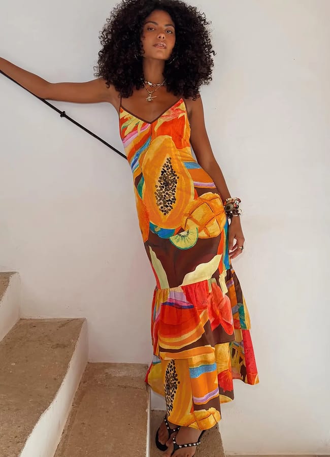 A woman with curly hair stands on a staircase, wearing a colourful, sleeveless maxi dress with bold, tropical fruit prints and black sandals. She leans against a white wall, looking at the camera.