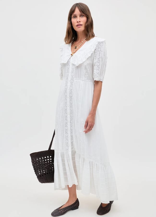 A woman wearing a white, lace-trimmed, midi-length dress with a large collar stands against a plain background. She holds a black woven tote bag and wears black woven flat shoes.