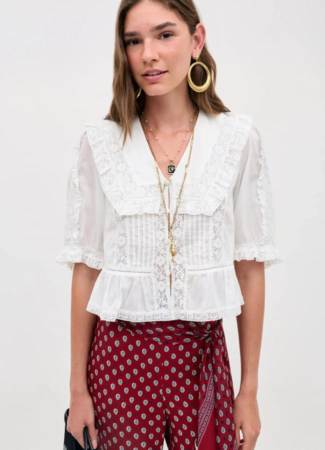 A woman wearing a sheer white lace summer top with ruffled trim, layered necklaces, large gold hoop earrings, and red patterned trousers stands against a plain light background.