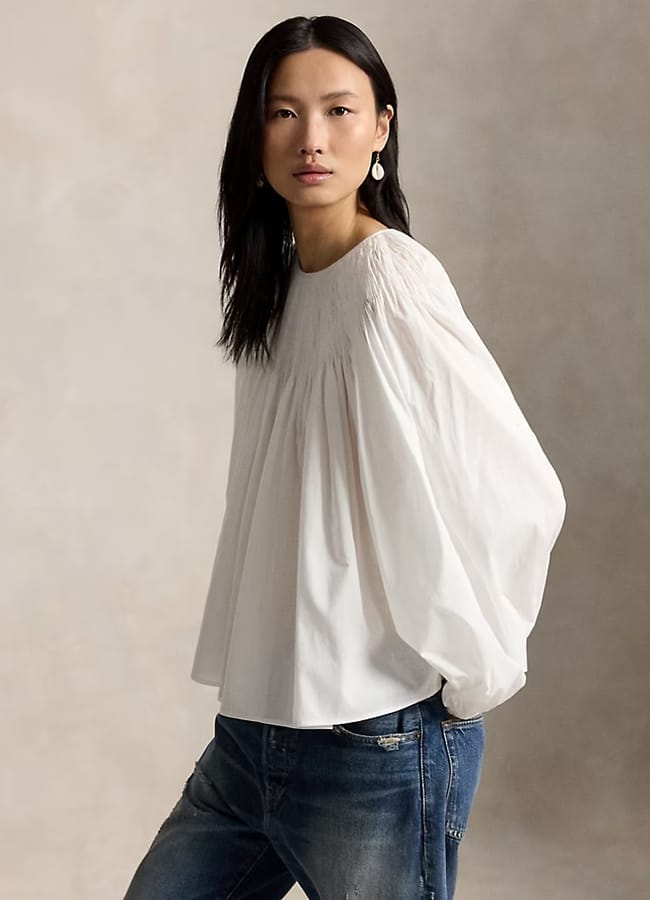 A woman with long black hair wears a loose, white, long-sleeved blouse—one of this season’s essential summer tops—and blue jeans. She stands against a neutral, softly lit background, calmly looking at the camera with a single dangling earring.