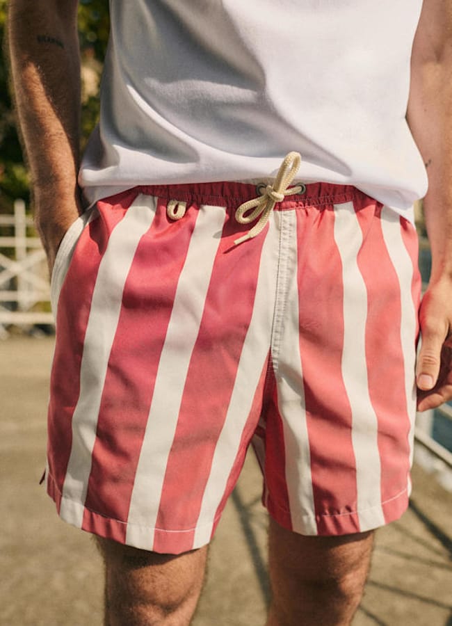 A person showcases men's holiday fashion in a white T-shirt and pink shorts with bold white vertical stripes, standing outdoors in sunlight. The shorts feature a beige drawstring at the waist.
