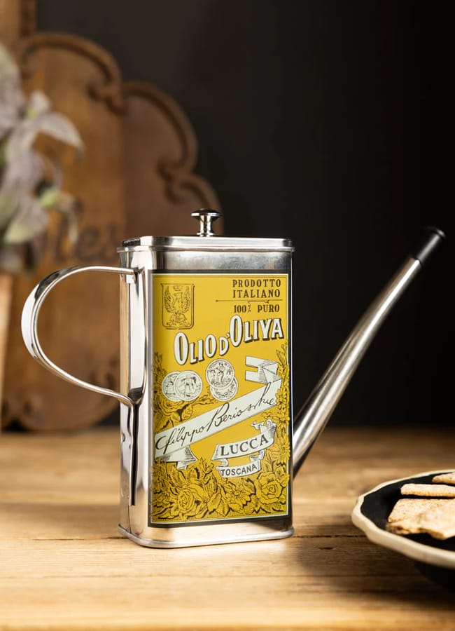 A vintage-style rectangular metal olive oil tin with a spout and handle, featuring a yellow Italian label, sits on a wooden table next to a plate of biscuits.