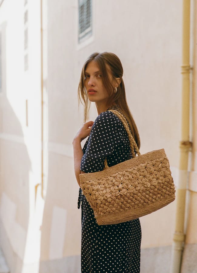 A woman with long brown hair wearing a black dress with white polka dots stands in a sunlit alleyway, looking over her shoulder. She has one of this season’s trendy basket bags slung over her arm.