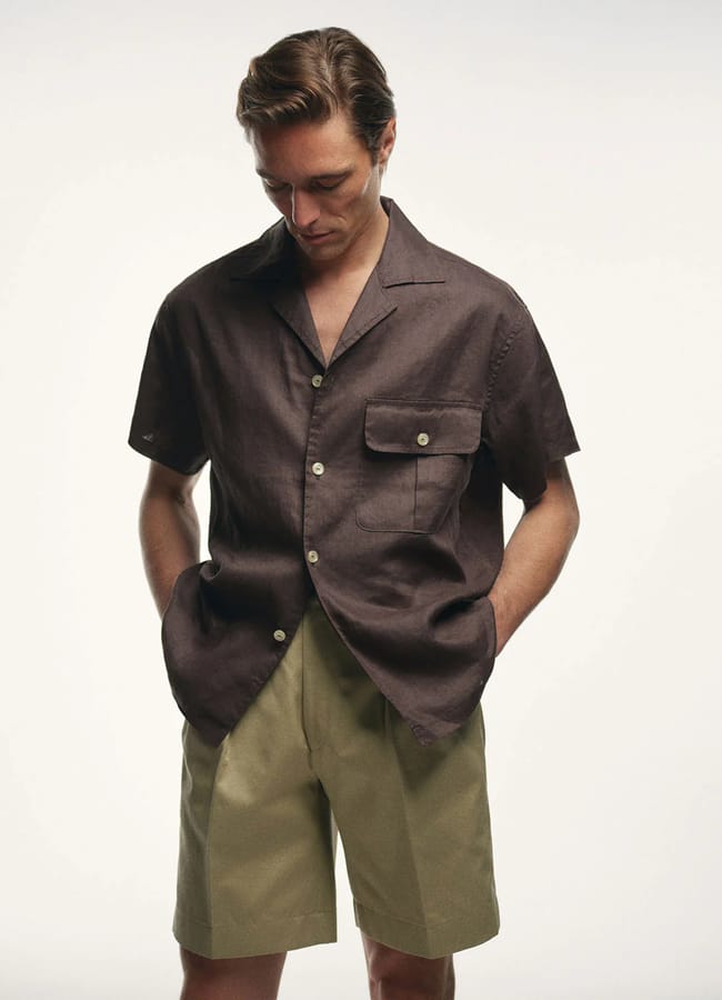 A man wearing a dark short-sleeve button-up shirt with a chest pocket and light khaki shorts stands with his hands in his pockets, looking down against a plain white background—a relaxed look perfect for men's holiday fashion.