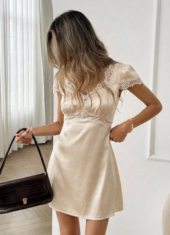 A woman with long, wavy hair wears a short, cream satin dress with lace details. She holds a dark brown handbag—an elegant pick for Fathers Day gifts 2026—and stands in a bright room with white curtains and a wooden floor.