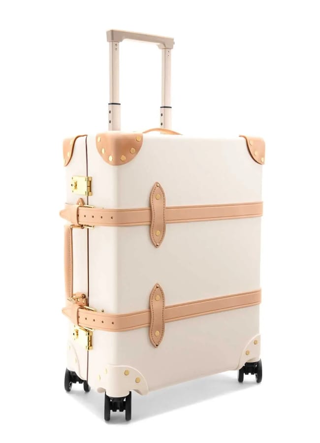 A vintage-style cream-coloured suitcase—evoking the charm of the best suitcase brands—features tan leather straps, gold fittings, a side handle, and four black spinner wheels. The telescopic handle is extended.