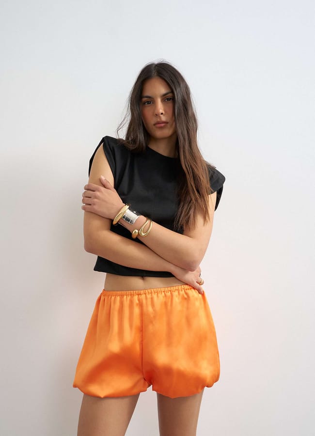 A woman with long brown hair stands against a plain white wall, wearing a black sleeveless crop top, bright orange satin bubble shorts, and several gold bangles on her wrist. She has a neutral expression and her arms are crossed.