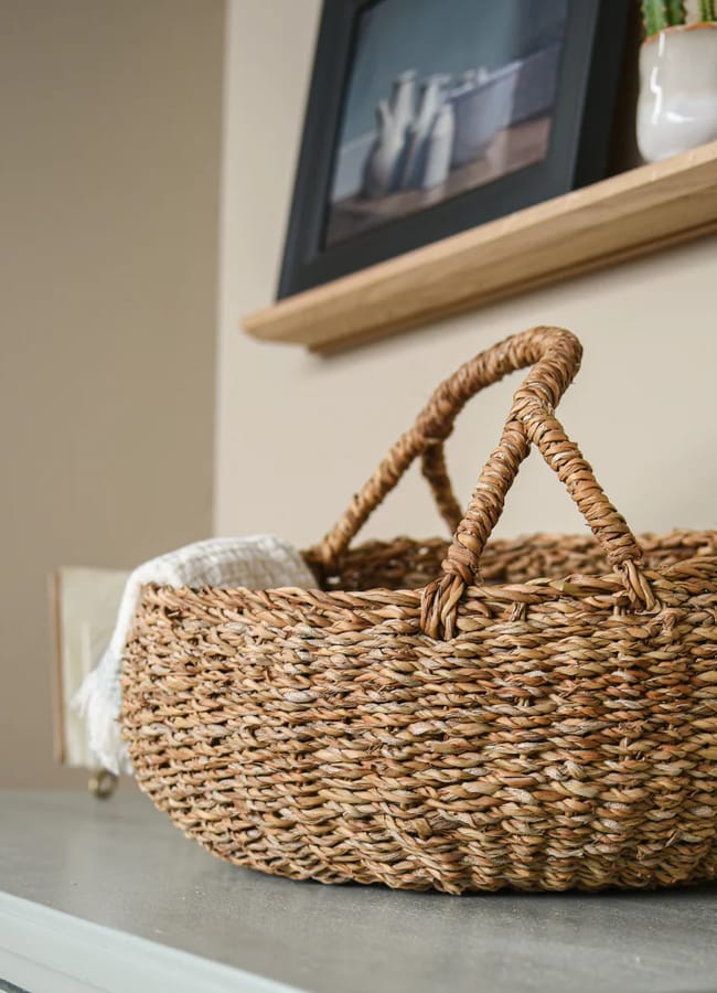 A woven wicker basket with two handles, perfect for picnic essentials, sits on a grey surface with a white cloth draped over one side. Above, a wooden shelf displays framed photos, a plant, and decorative items against a beige wall.