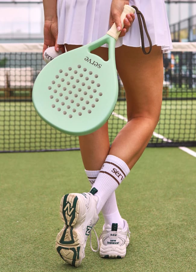 A person in a white tennis skirt and trainers stands on a court, holding a light green paddle with the brand name Serve. Their legs are crossed, showing off tall white socks—a stylish choice for cute padel outfits or trendy padel clothing.