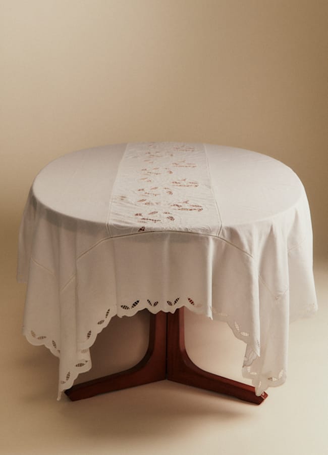 A round wooden table covered with a white tablecloth and a decorative lace table runner, set against a plain beige background.