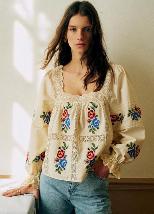 A woman with long brown hair wears a cream-coloured blouse with floral embroidery and lace details, paired with blue jeans. She stands indoors in soft natural light, resting one hand on a table.