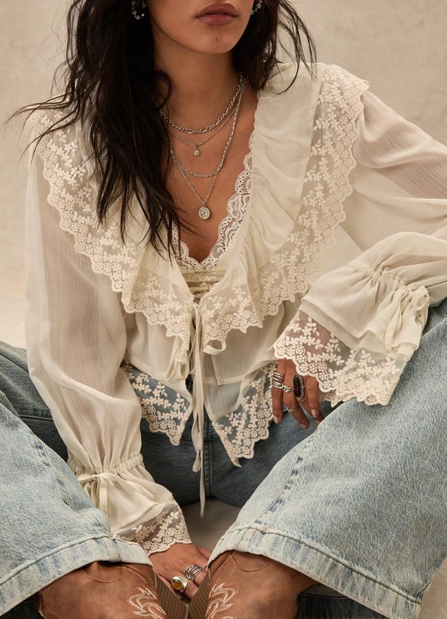 A woman wearing a sheer, frilled white summer top with lace details, layered necklaces, rings, and light blue jeans. She is seated with her hands resting on her legs; her face is partially out of frame.