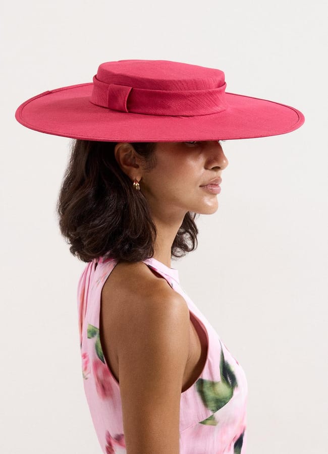 A woman with shoulder-length dark hair wears a wide-brimmed, bright pink hat and a sleeveless floral dress—serving a hot take in style against a plain white background.