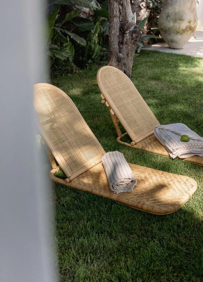 Two woven rattan loungers with striped towels, a lime, and picnic essentials rest on green grass in a garden, surrounded by leafy plants and trees.