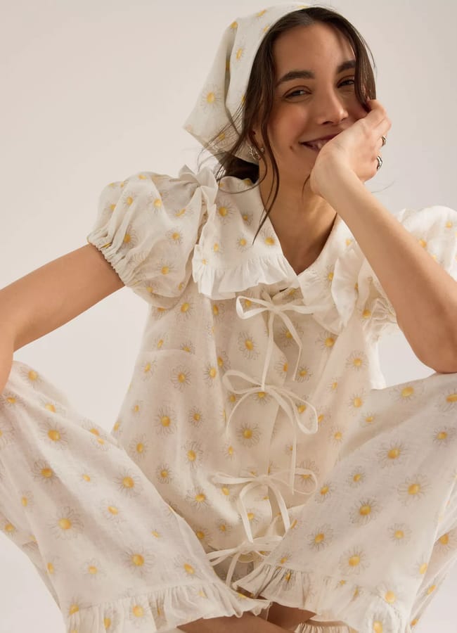 A smiling woman wearing a matching cream-coloured pyjama set with a daisy print and a headscarf sits cross-legged. The outfit features puffed sleeves, lace-up front, ruffled edges, and a wide collar.