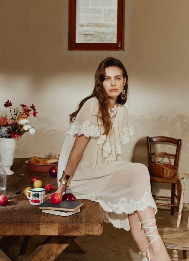 A woman in a cream lace dress sits on a rustic wooden table with fruit, flowers, books, and a mug—perfect inspiration for unique Fathers’ Day gifts 2026. She looks at the camera, with a neutral wall, window, and wooden chair behind her.