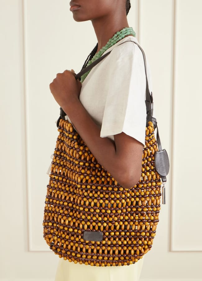 A person wearing a white short-sleeve top carries a large beaded bag with leather handles over their shoulder, standing against a cream-coloured wall.