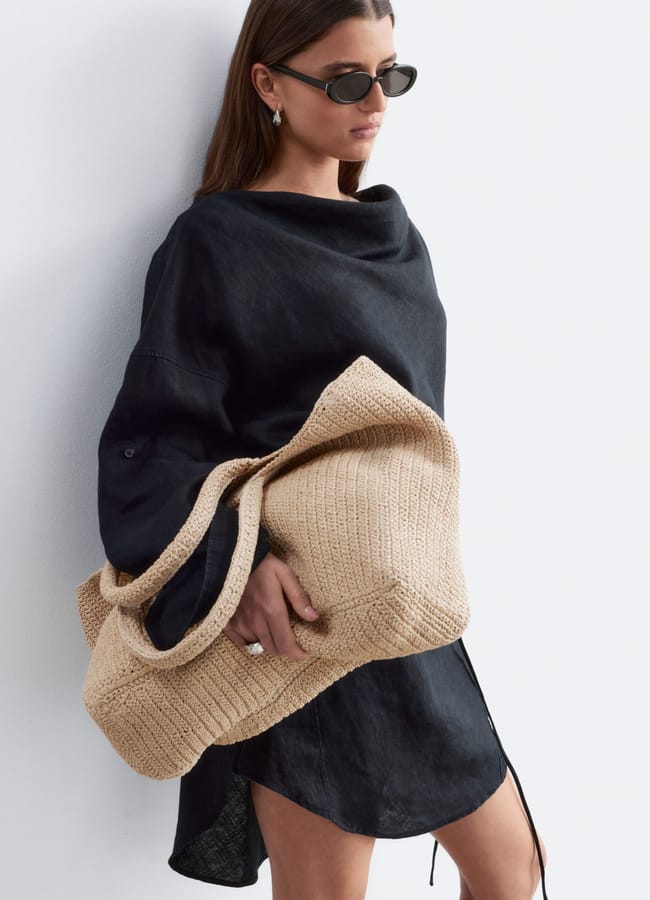 Woman wearing a loose black dress and dark sunglasses, holding a large beige woven basket bag, stands against a plain white background.