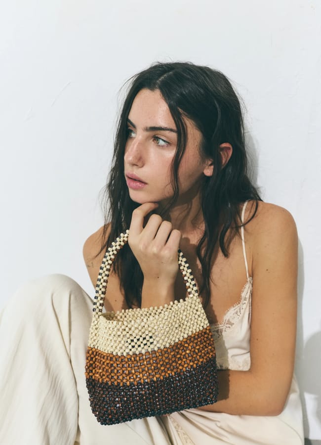 A woman with long dark hair in a cream-coloured dress sits by a white wall, holding a chic beaded bag striped in cream, brown, and dark brown. She gazes thoughtfully to the side.