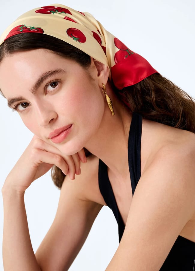 Woman with long brown hair wears a cream-coloured headscarf decorated with playful fruit prints of red tomatoes, gold earring, and a black halter top, looking at the camera with her chin resting on her hand.